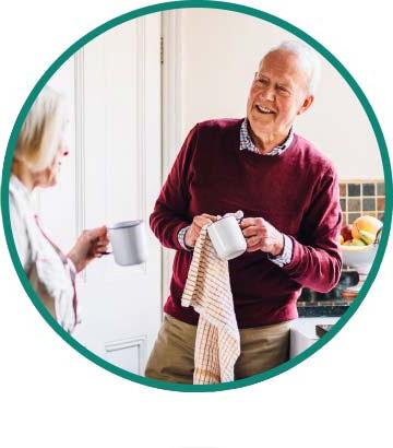 An elderly man cheerfully dries dishes while talking with his wife